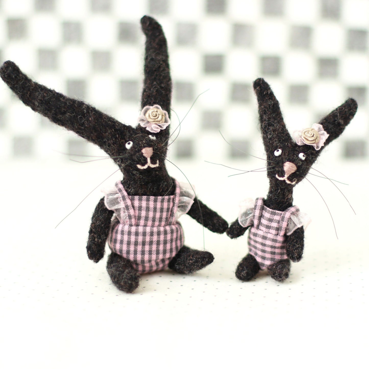andmade dark brown wool bunny collectible plush set — miniature rabbits in pink checked outfits with floral headpieces, sitting and standing together.