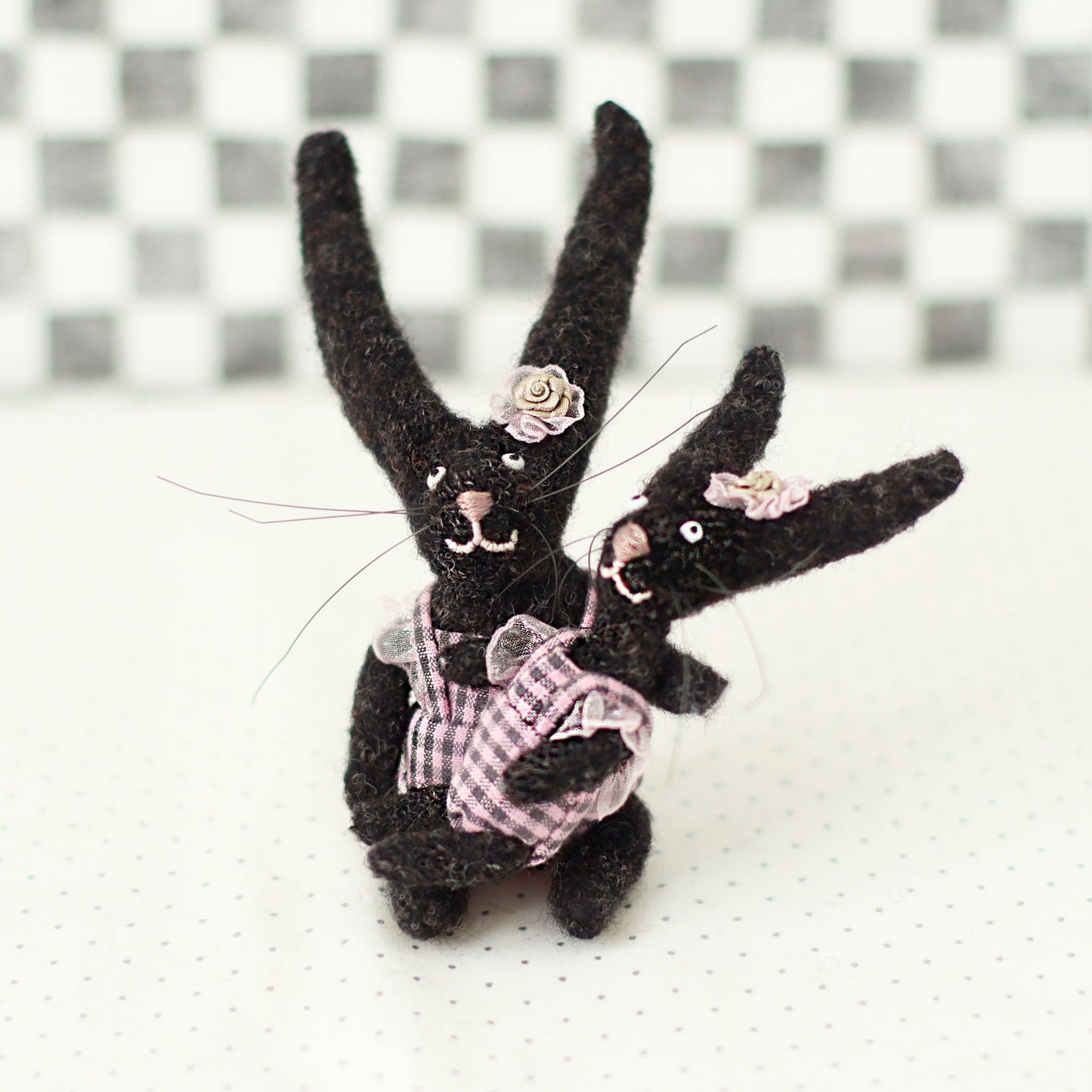 Handmade dark brown wool bunny collectible plush set — miniature rabbits in pink checked outfits with floral headpieces, hugging in a seated pose.