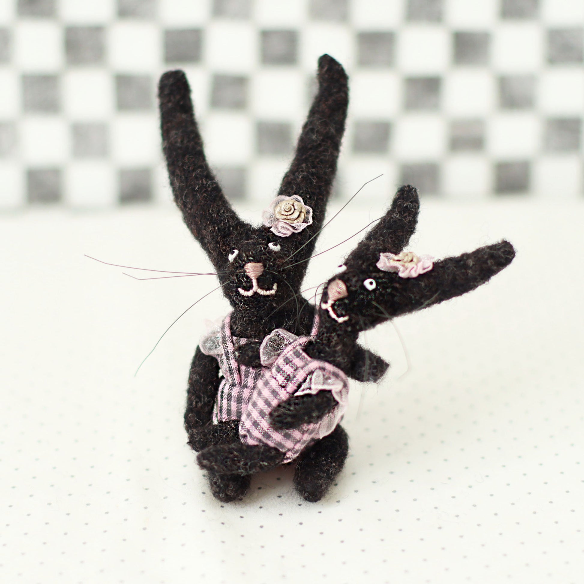 Handmade dark brown wool bunny collectible plush set — miniature rabbits in pink checked outfits with floral headpieces, hugging in a seated pose.