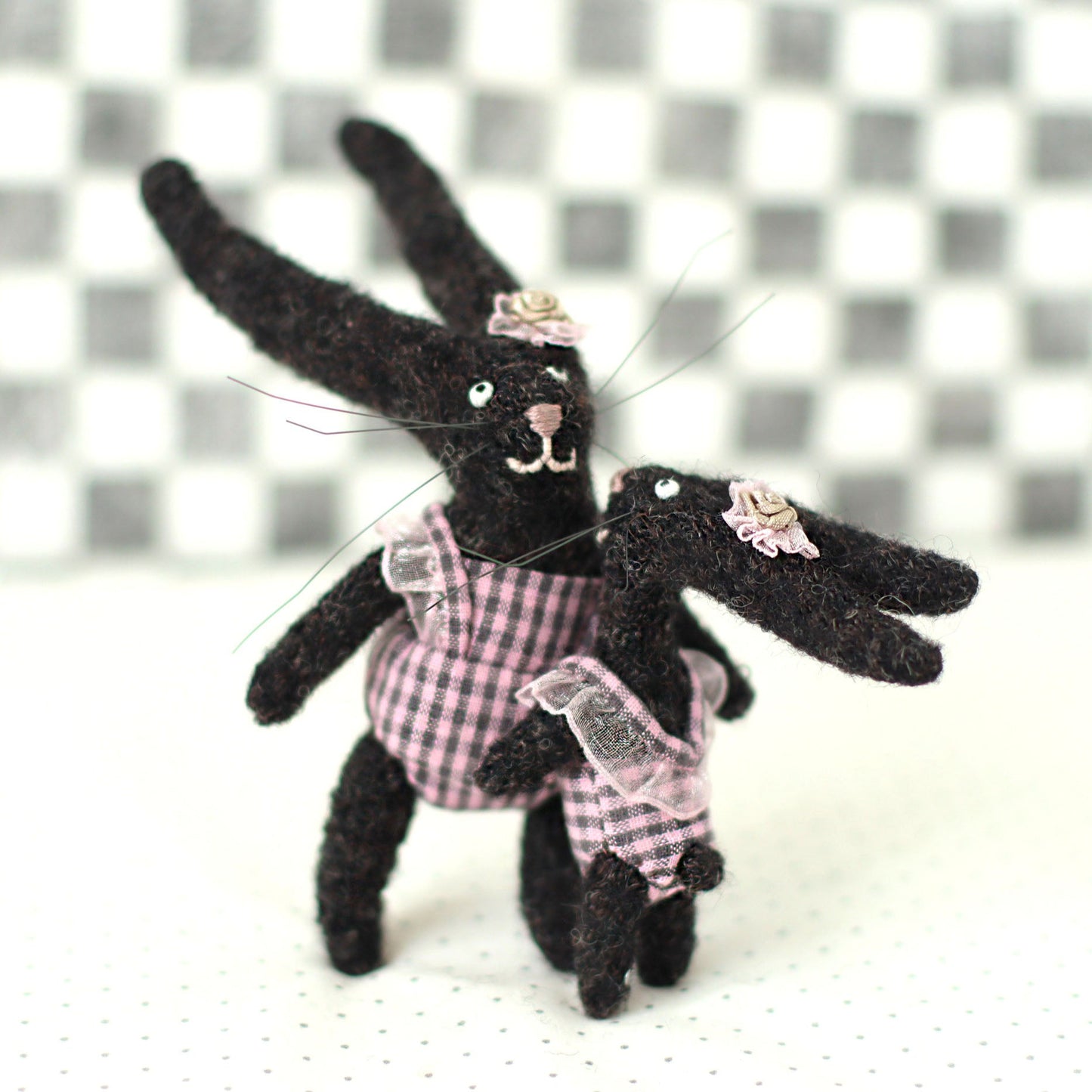 Handmade dark brown wool bunny collectible plush set — miniature rabbits in pink checked outfits with floral headpieces, standing closely together in a playful pose.