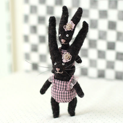 Handmade dark brown wool bunny collectible plush set — miniature rabbits in pink checked outfits with floral headpieces, posed with the baby bunny riding on the adult’s shoulders.