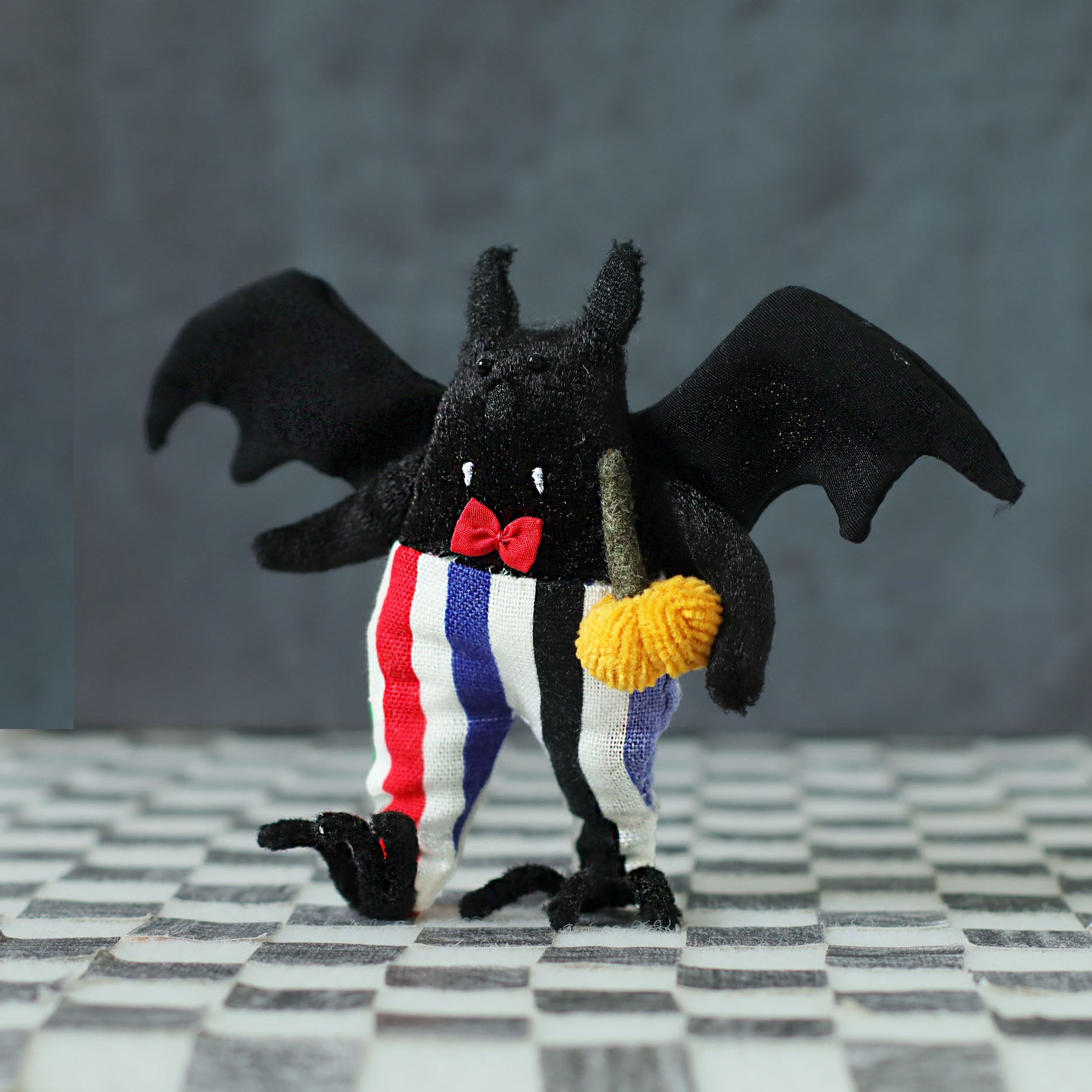Handmade bat plush toy — soft black fabric bat with wings, red bow tie, striped trousers, and a miniature pumpkin, posed on a checkered floor as a collectible art doll