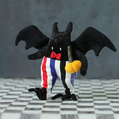 Handmade black bat plush toy with wide wings, striped red-blue-white pants, a red bow tie, and a small yellow pumpkin, standing on a checkered surface