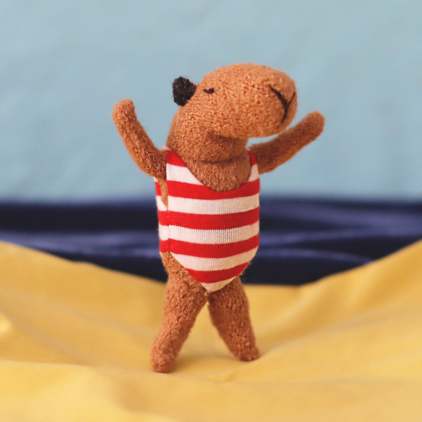 Miniature handmade capybara doll in a red-and-white striped swimsuit, standing with raised arms on yellow “sand” fabric with a blue sea-and-sky background