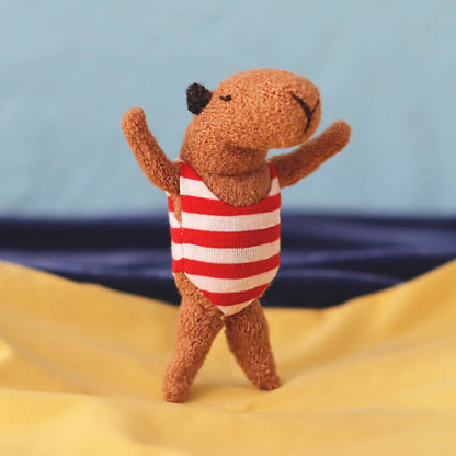 Miniature handmade capybara doll in a red-and-white striped swimsuit, standing with raised arms on yellow “sand” fabric with a blue sea-and-sky background