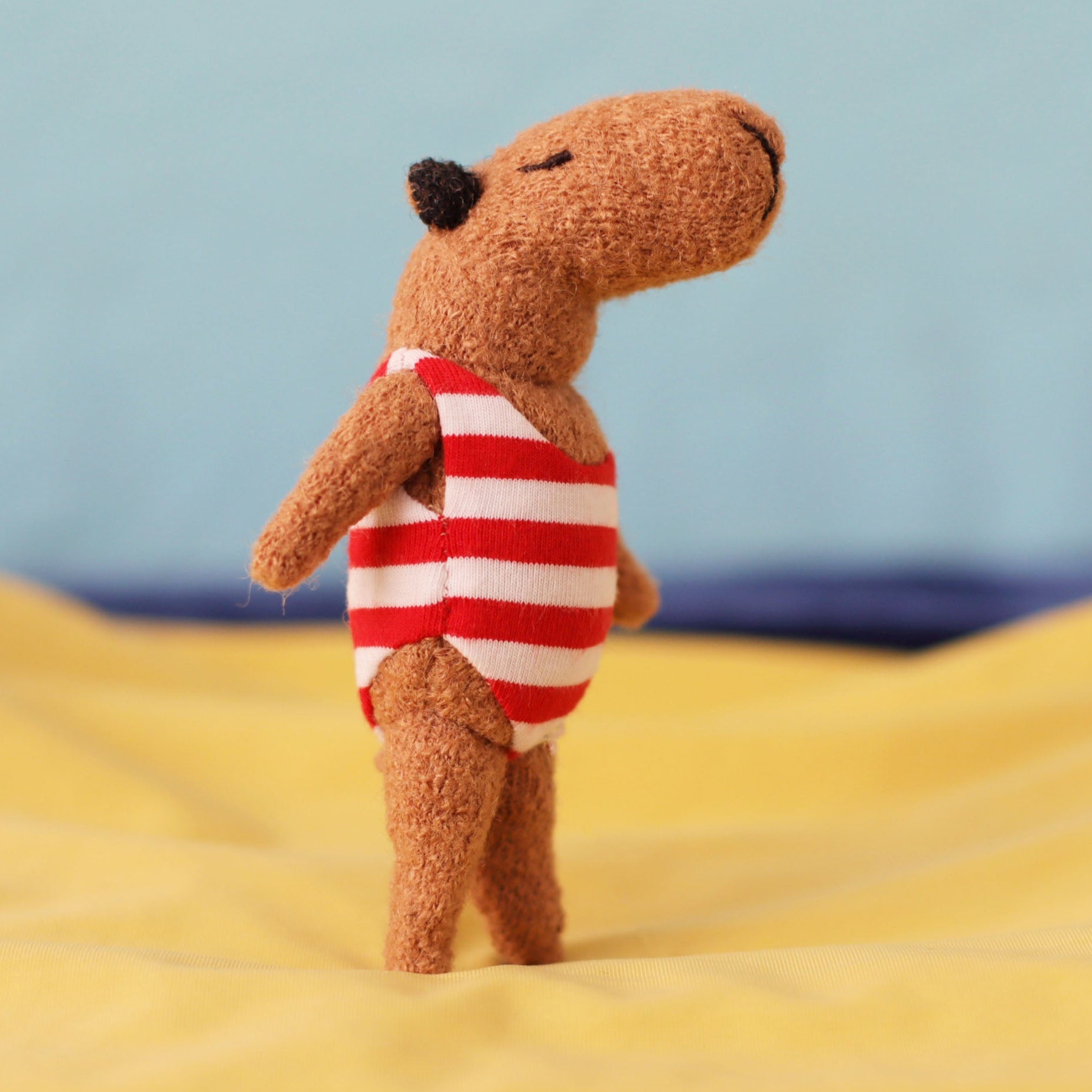 Handmade miniature capybara doll standing upright, wearing a red-and-white striped swimsuit, posed on soft yellow fabric with a blue background