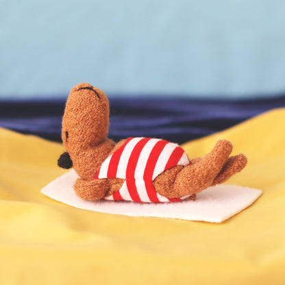 Miniature handmade capybara doll in a red-and-white striped swimsuit, lying on a tiny white towel and sunbathing on yellow “sand” fabric with a blue sea-and-sky background
