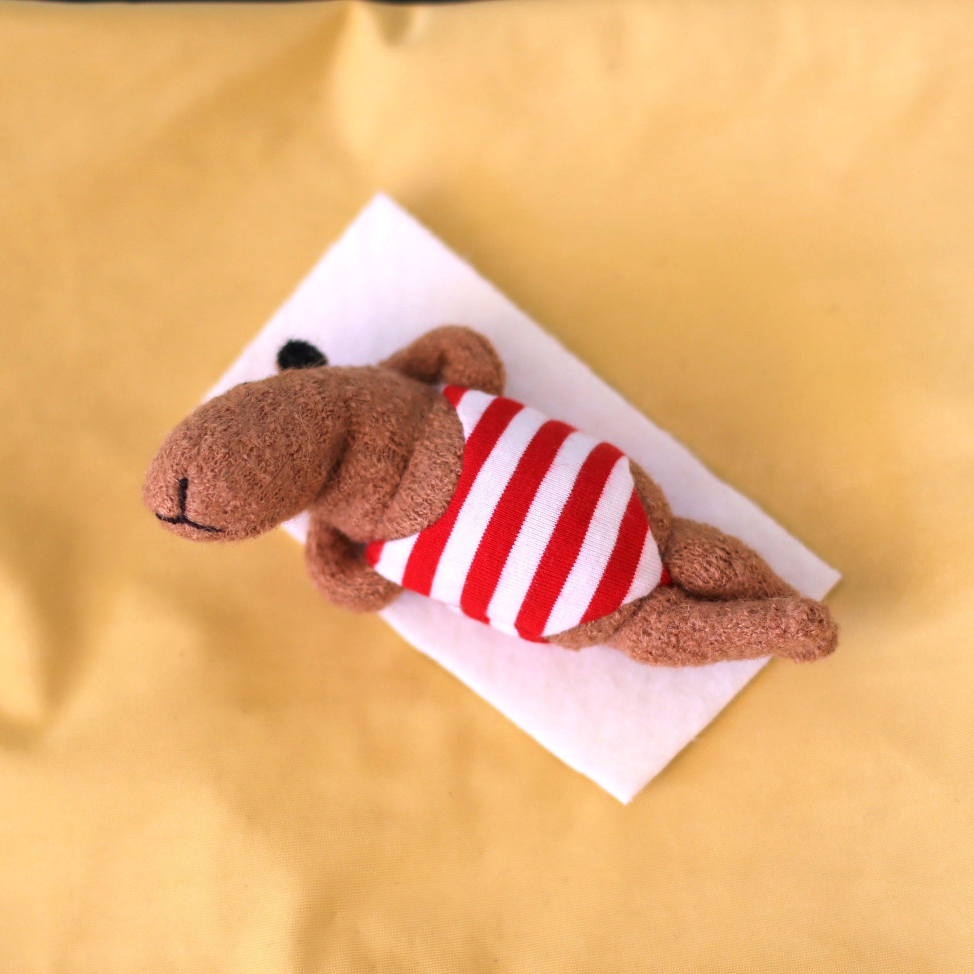 Handmade miniature capybara doll wearing a red-and-white striped swimsuit, lying on a small white towel on yellow “sand” fabric, posed as if sunbathing