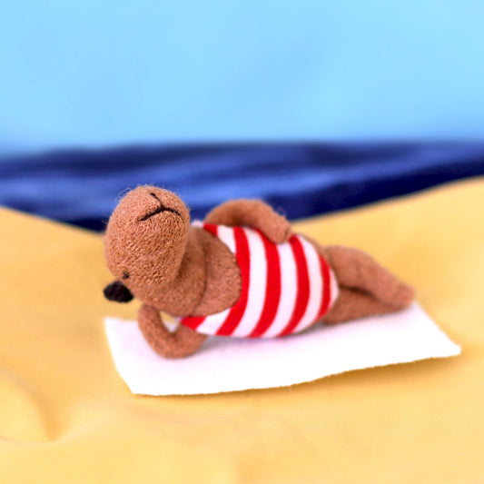 Handmade miniature capybara doll lounging on a small white towel, wearing a red-and-white striped swimsuit, posed on yellow fabric resembling sand with a blue “sea and sky” background