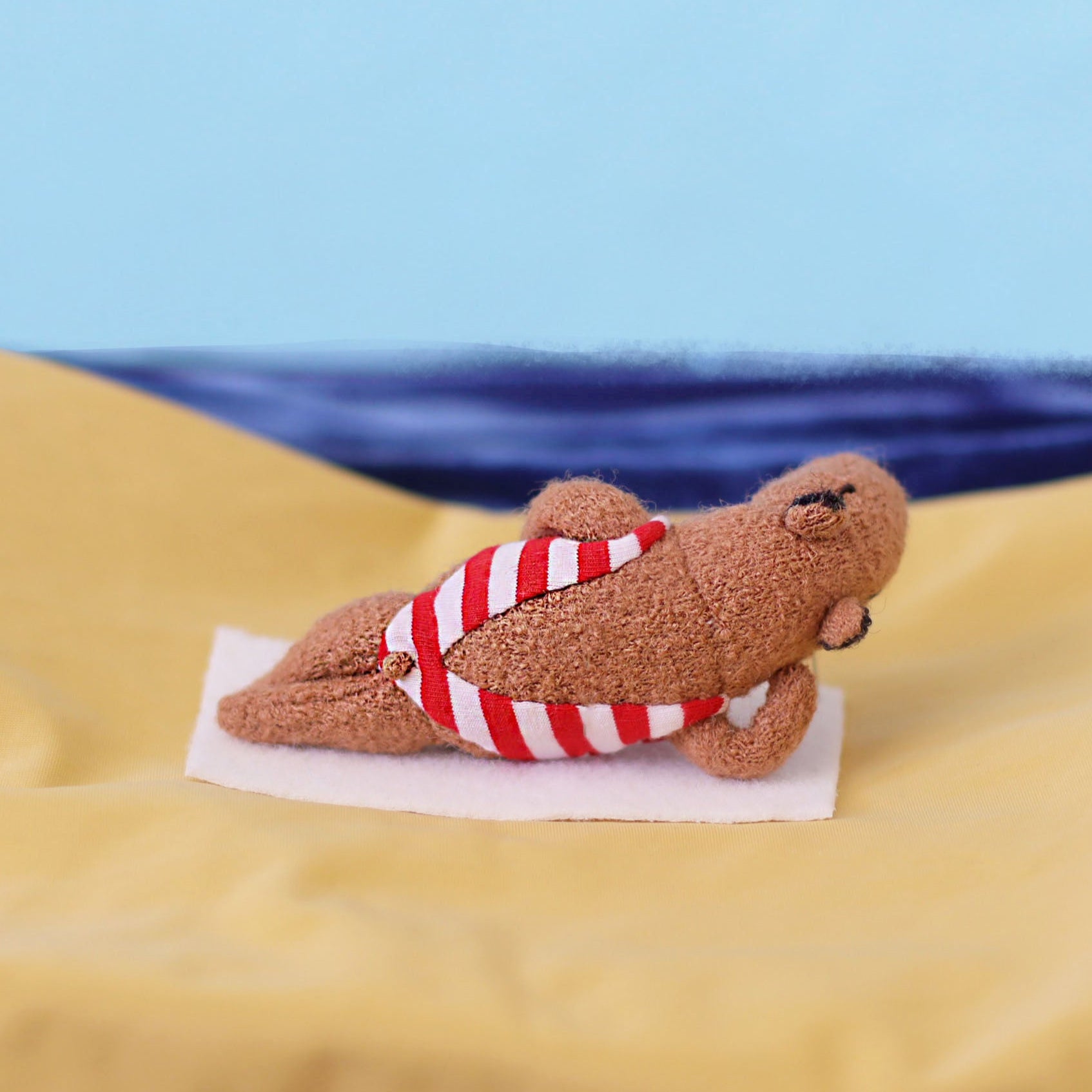 Handmade miniature capybara doll lying on its back on a small white towel, wearing a red-and-white striped swimsuit, posed on yellow fabric as “sand” with a blue sea-and-sky background