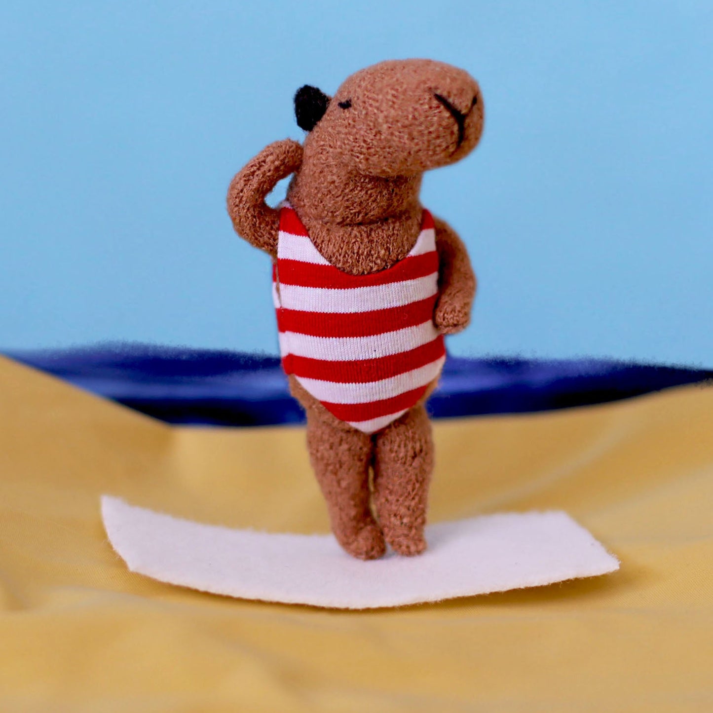 Handmade miniature capybara doll standing on a small white towel, wearing a red-and-white striped swimsuit, posed on yellow “sand” fabric with a blue sea-and-sky backdrop