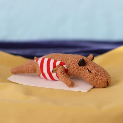 Handmade miniature capybara doll lying face down on a small white towel, wearing a red-and-white striped swimsuit, posed on yellow “sand” fabric with a blue sea-and-sky background