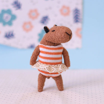 Handmade miniature capybara doll wearing an orange striped dress with a lace trim, standing against a soft blue and floral background