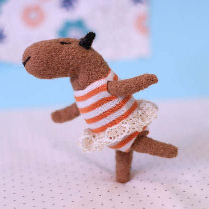 Miniature handmade capybara doll balancing on one leg, wearing an orange-striped dress with a lace hem against a soft blue and patterned background