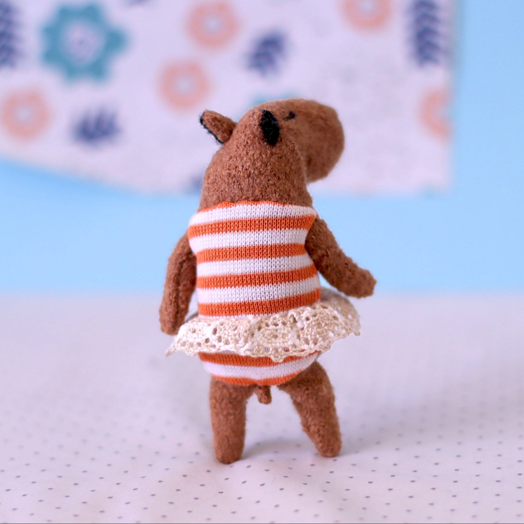 Miniature handmade capybara doll shown from the back, wearing an orange-striped dress with a lace hem, standing on a dotted surface with a pastel blue and floral background