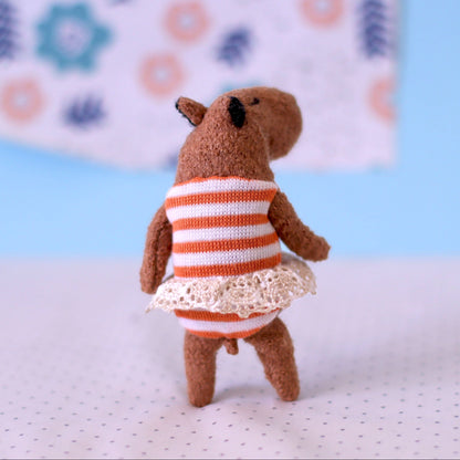 Miniature handmade capybara doll shown from the back, wearing an orange-striped dress with a lace hem, standing on a dotted surface with a pastel blue and floral background