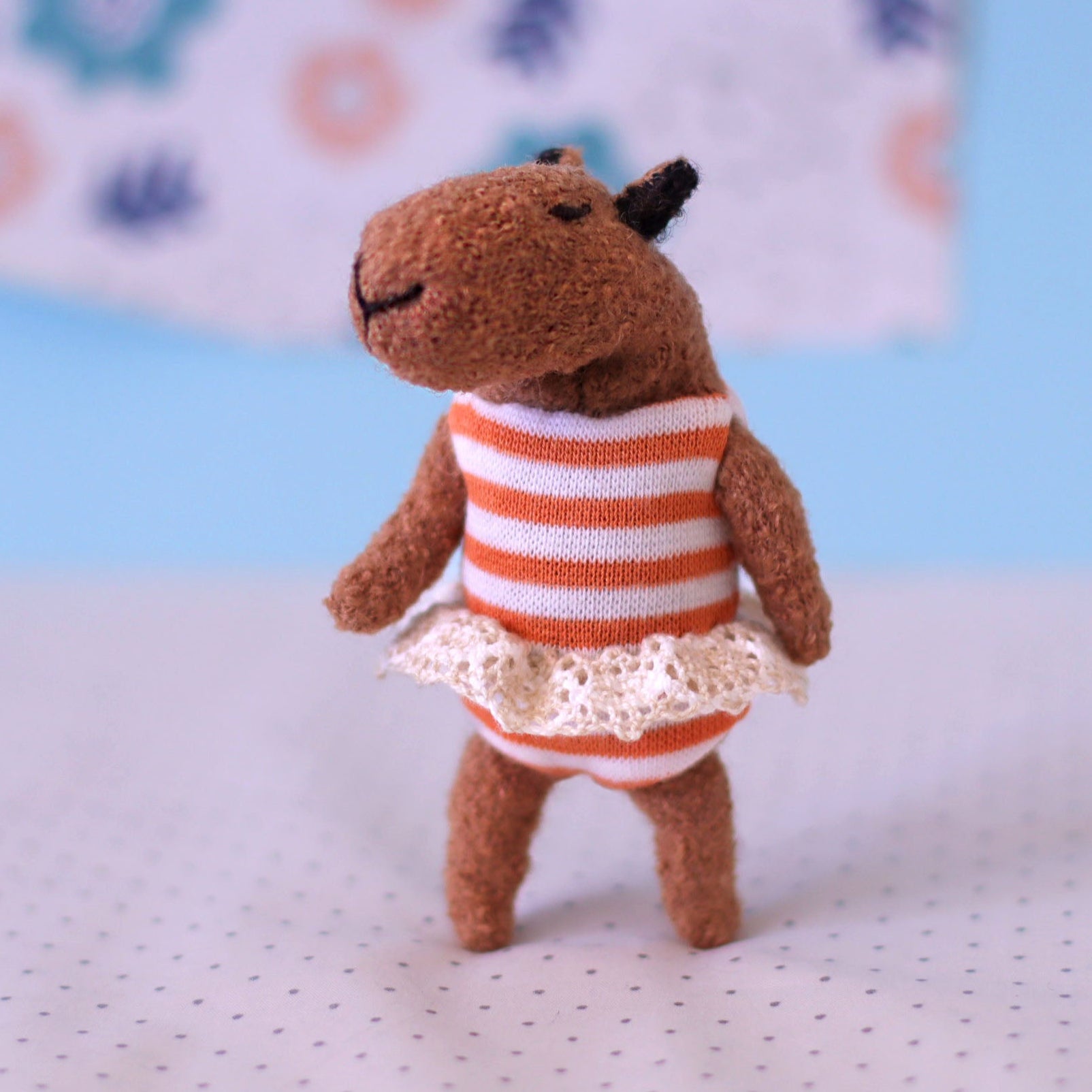 iny handmade capybara doll in an orange-striped dress with a lace trim, standing on a dotted surface with a pastel blue and floral background