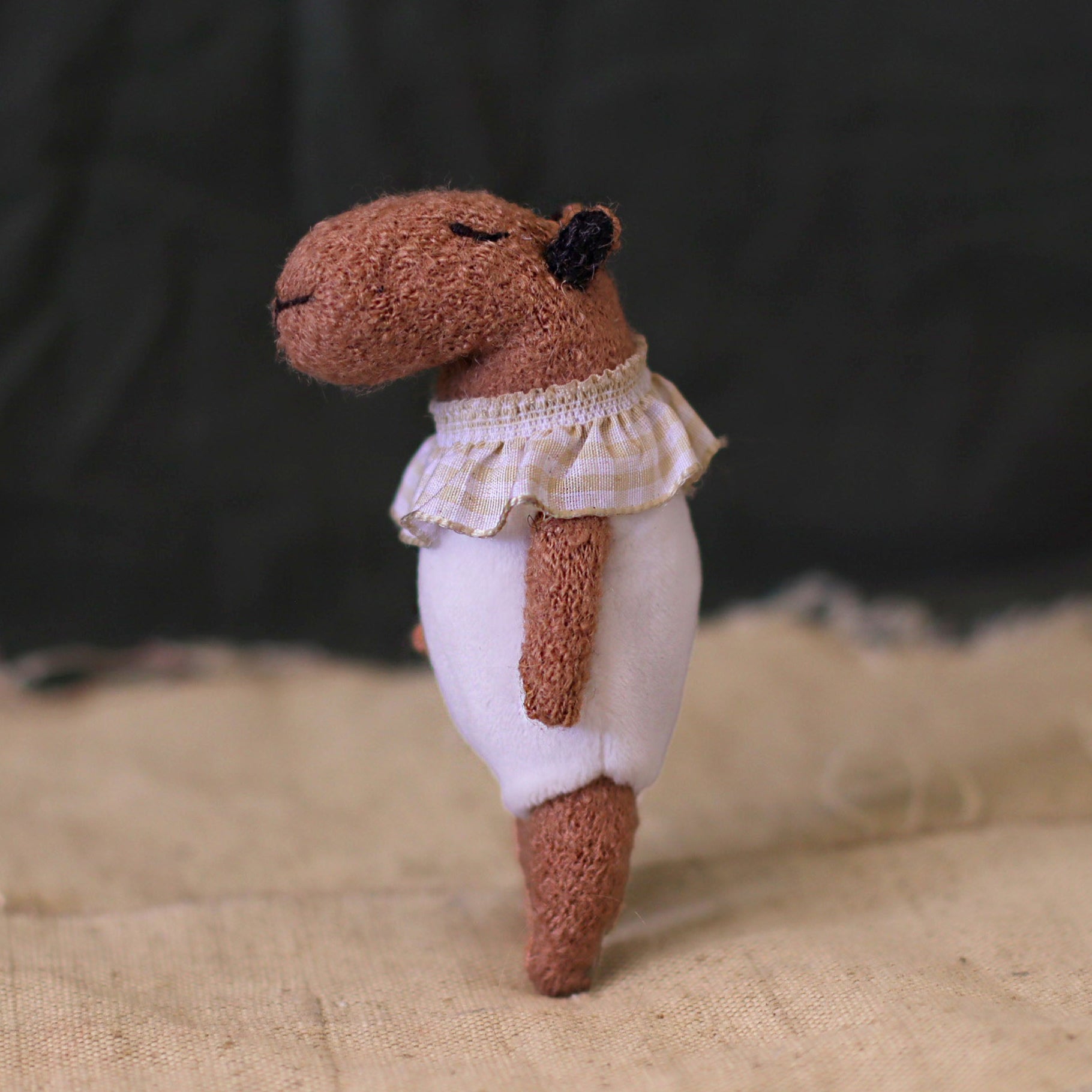 Side view of a handmade miniature capybara doll in a white romper with a ruffled checked collar, standing on beige fabric with a peaceful closed-eye expression