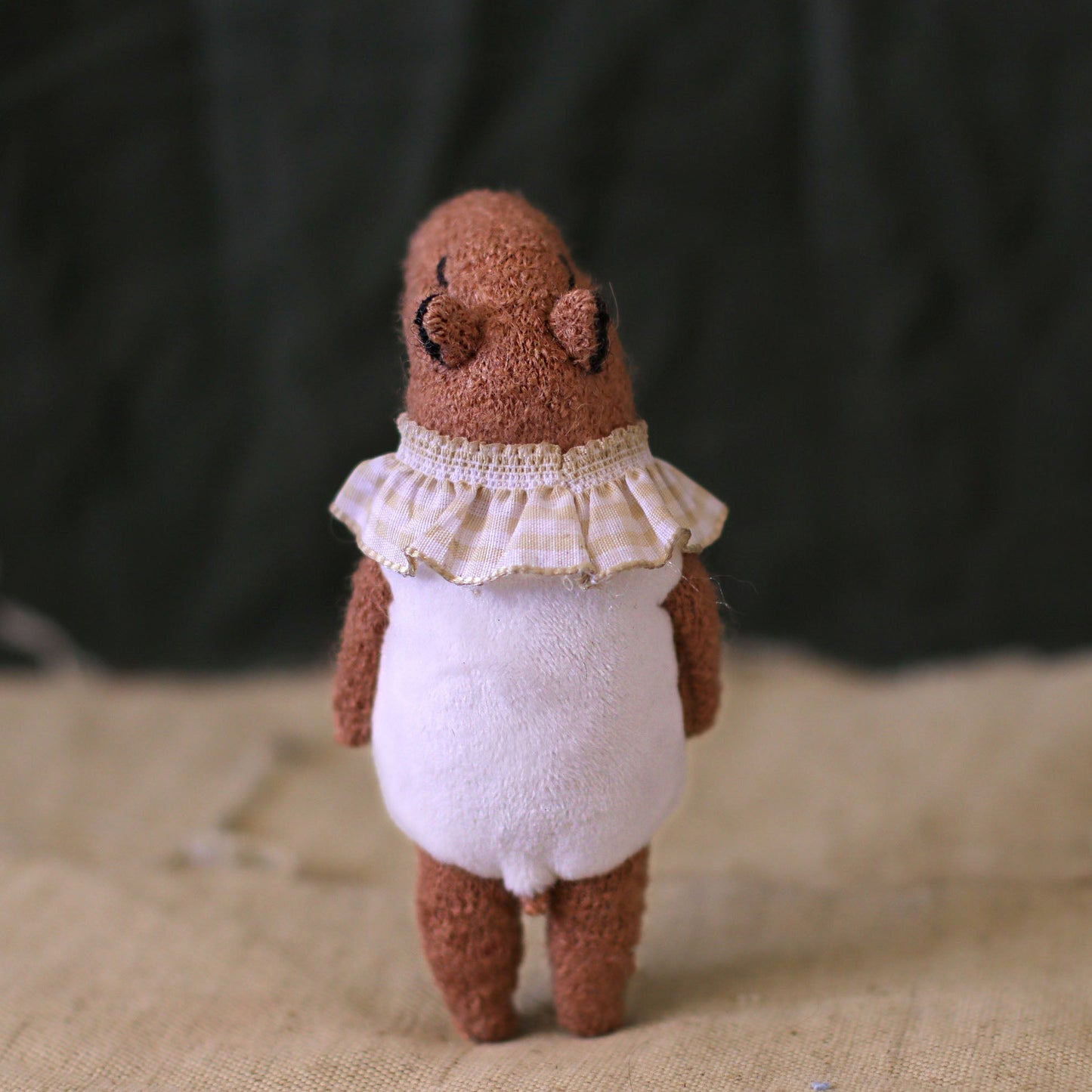Back view of a handmade capybara doll wearing a white romper and a frilled gingham collar, standing on a textured fabric background