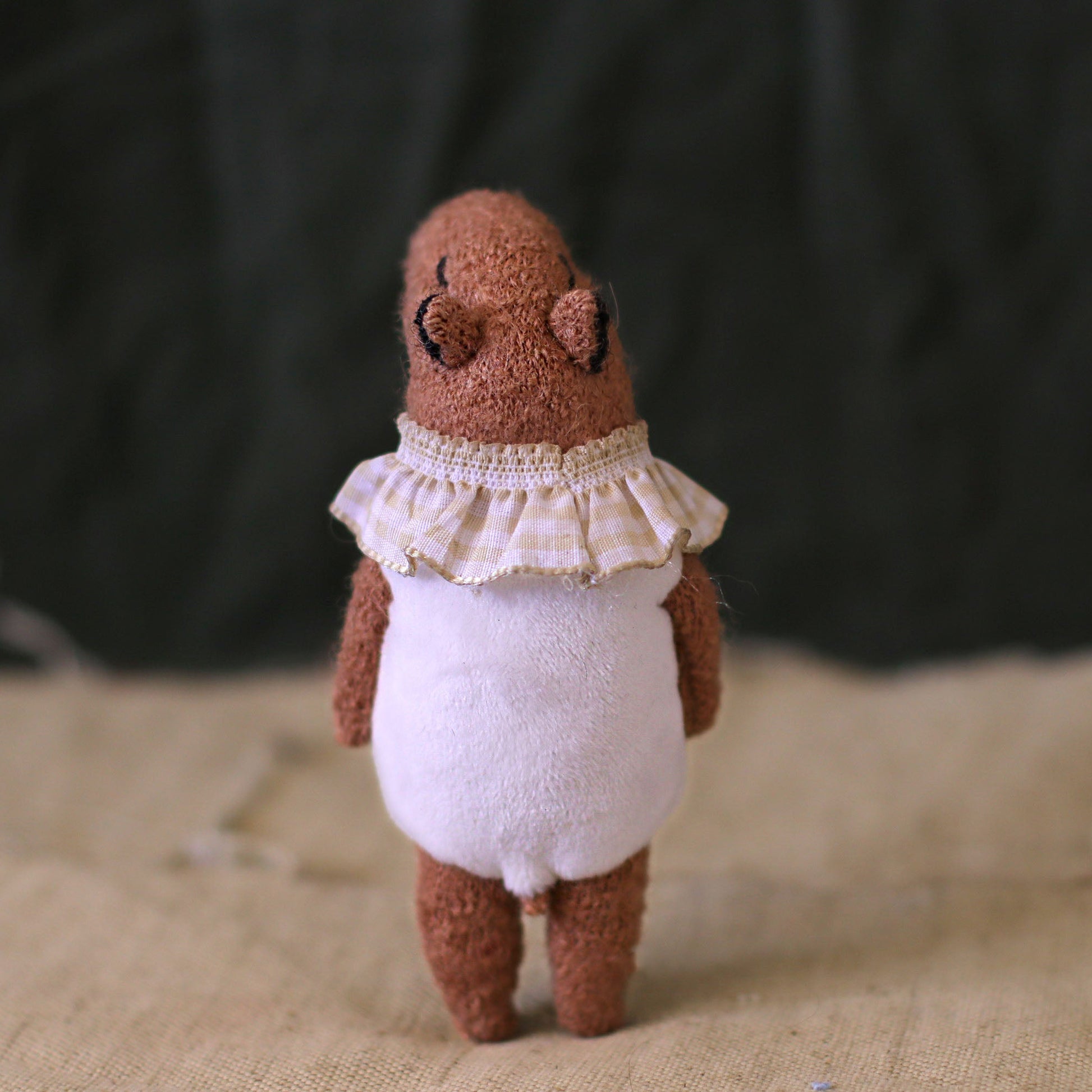 Back view of a handmade capybara doll wearing a white romper and a frilled gingham collar, standing on a textured fabric background