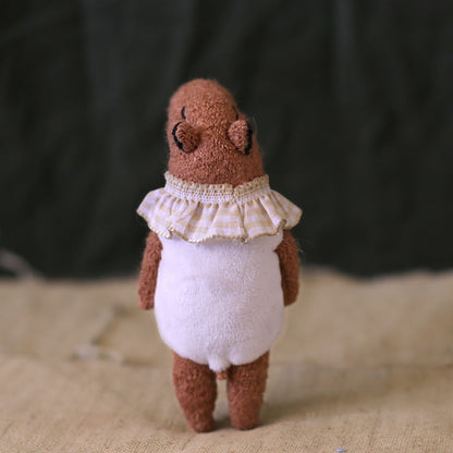 Back view of a handmade capybara doll wearing a white romper and a frilled gingham collar, standing on a textured fabric background
