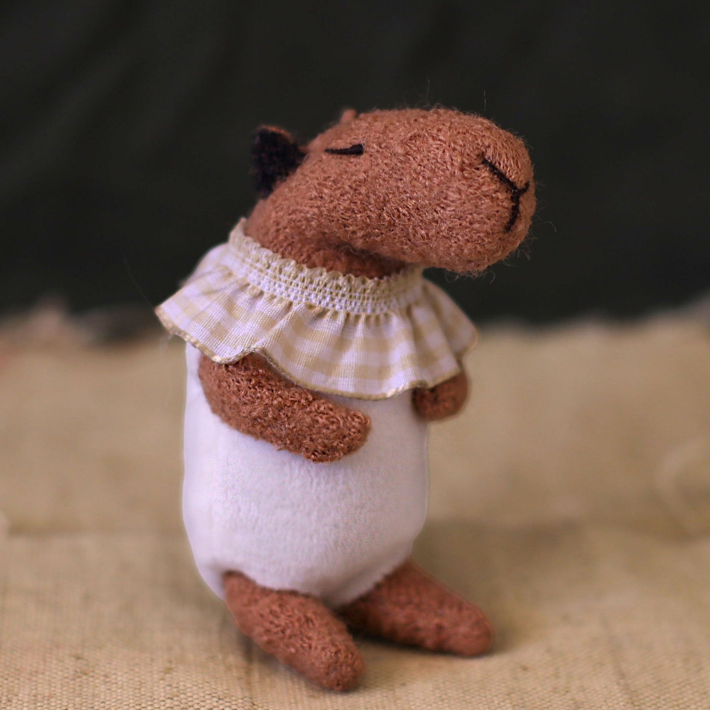 Handmade capybara doll sitting on a rustic fabric surface, wearing a white romper and a beige gingham ruffled collar, with eyes closed in a peaceful, content expression