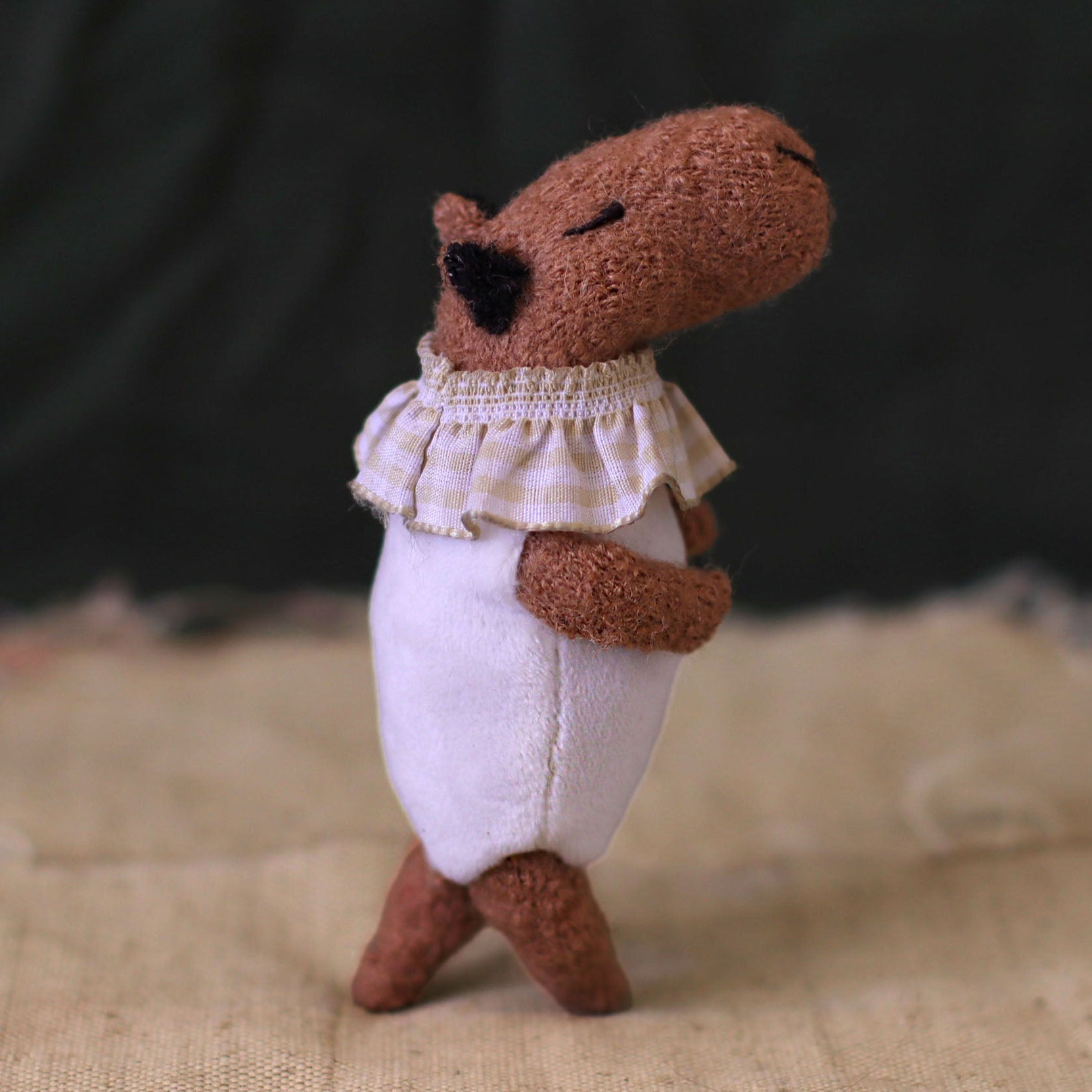 Handmade capybara doll in a white romper and ruffled gingham collar, shown in a side view as if walking on rustic fabric