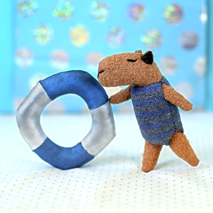 Miniature handmade capybara doll in a blue striped swimsuit leaning on a small blue-and-white swim ring