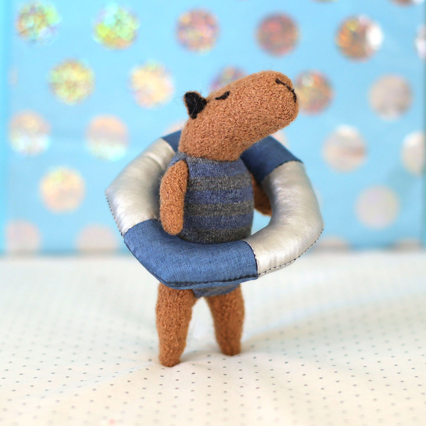 Miniature handmade capybara doll in a blue striped swimsuit standing inside a small blue-and-white float ring