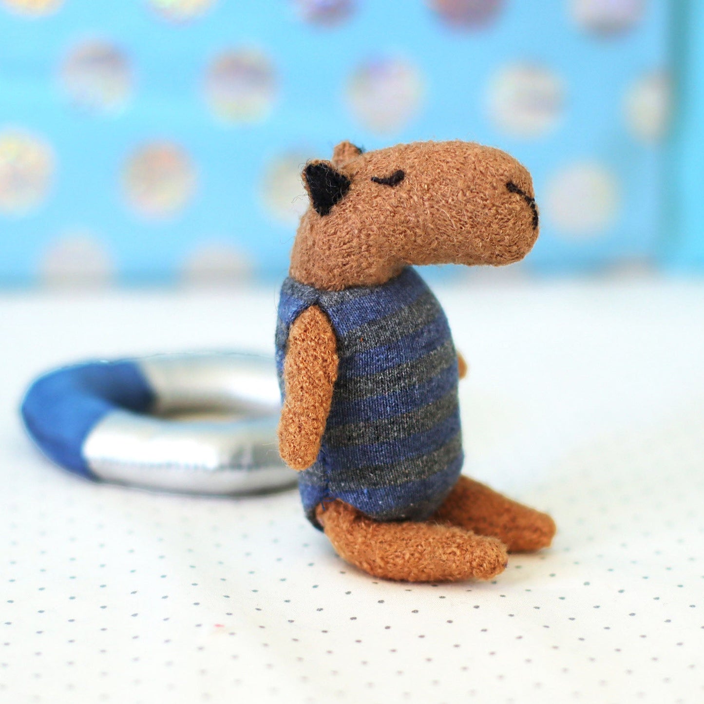 Miniature handmade capybara doll in a blue striped swimsuit sitting on a dotted background with a small swim ring behind it