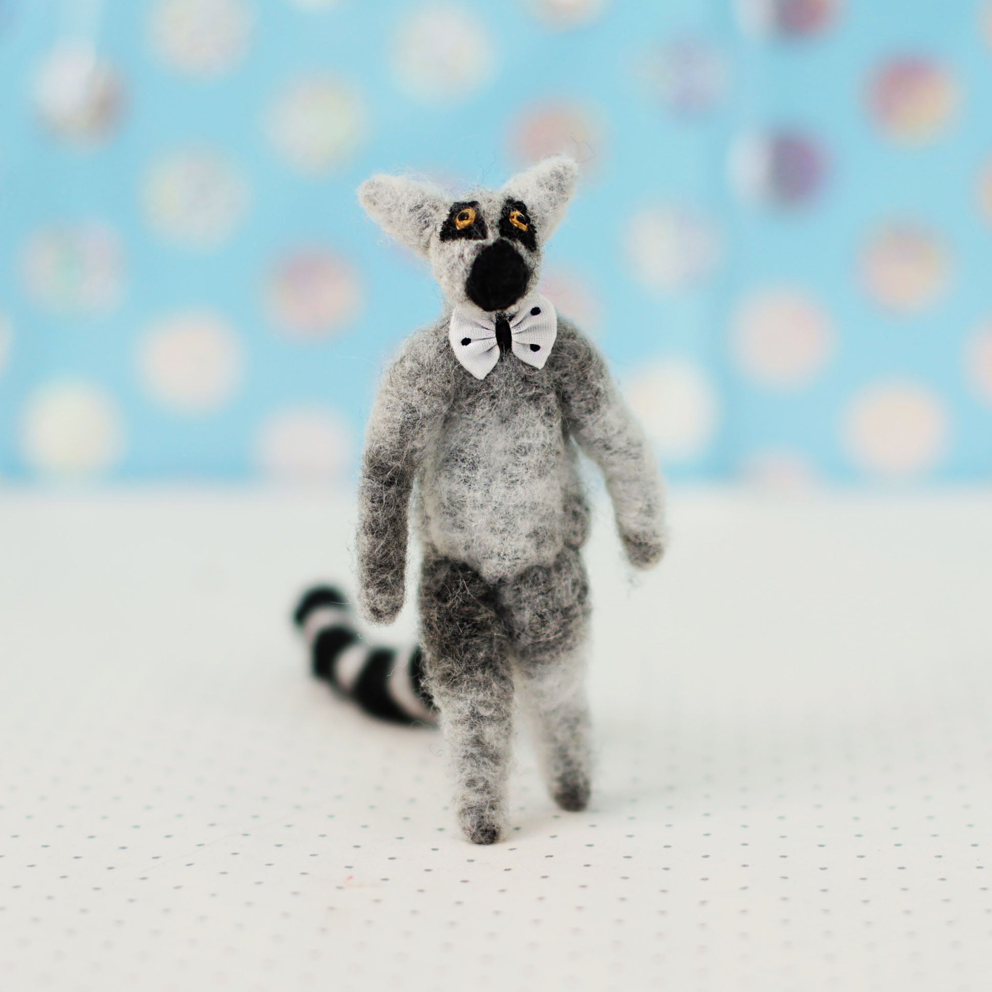 Hand-sewn miniature ring-tailed lemur plush toy with a striped tail and tiny bow tie, standing upright against a light blue background.