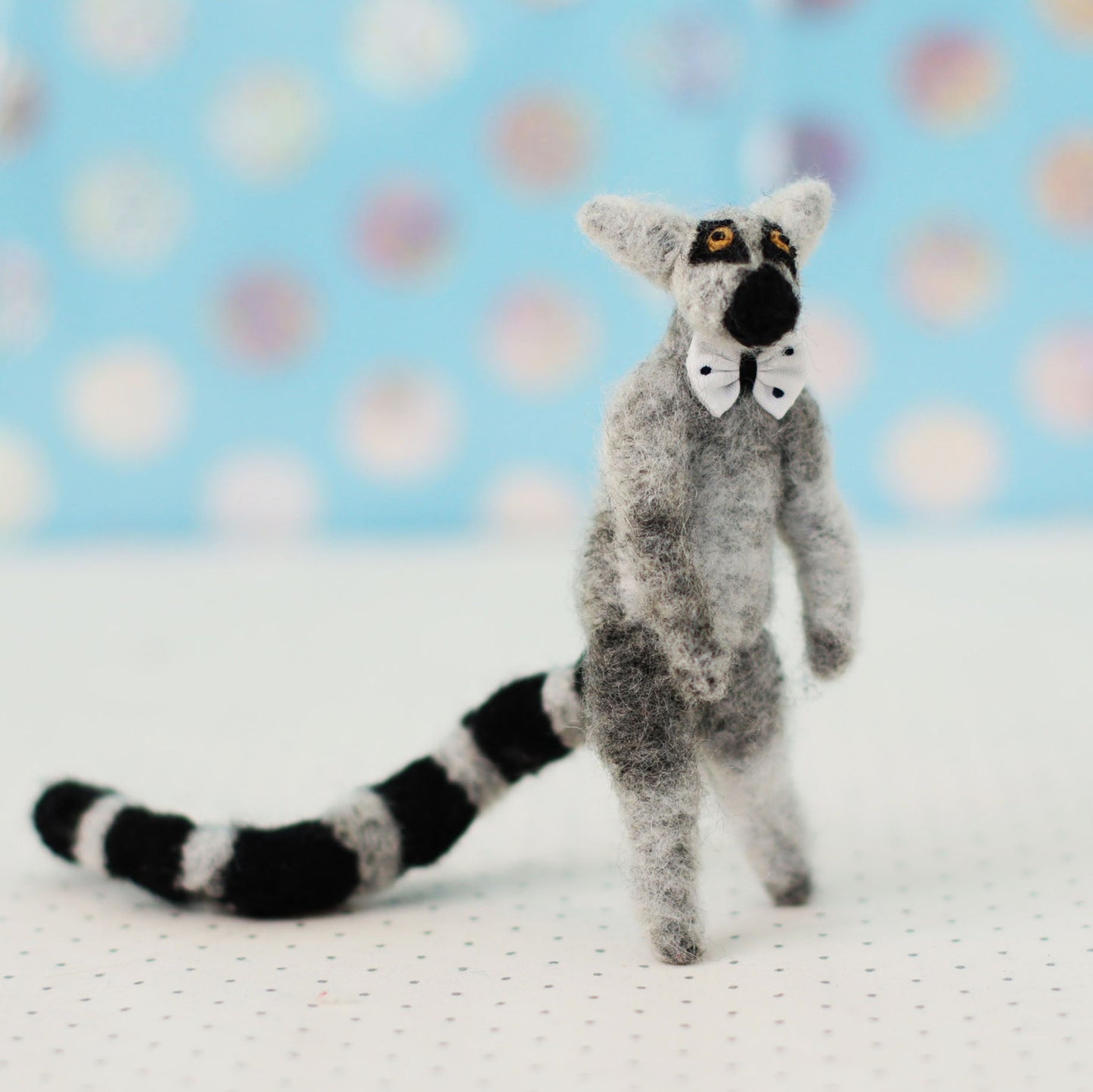 Hand-sewn miniature ring-tailed lemur plush toy with a black-and-white striped tail and small bow tie, standing upright against a soft blue background.
